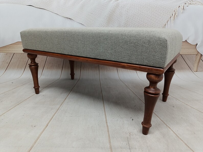 May include: A light grey upholstered bench with a wooden frame and turned legs. The bench has a simple, elegant design and would be a great addition to any home.