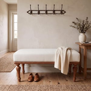 May include: A rectangular upholstered bench with a cream-coloured fabric seat and turned wooden legs. A cream throw blanket is draped over one side of the bench. A pair of brown leather loafers sits on the floor beneath the bench.