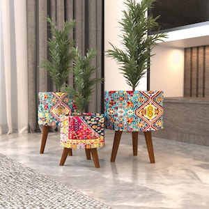 Puede incluir: Tres macetas cilíndricas con tres patas de madera cada una, con un vibrante patrón geométrico multicolor en tonos azul, rojo, amarillo y blanco. Cada maceta contiene una planta verde con hojas finas y puntiagudas de diferentes alturas.