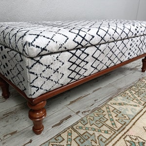 May include: A white and black patterned storage ottoman with a wooden frame and turned legs. The ottoman is sitting on a light wood floor.