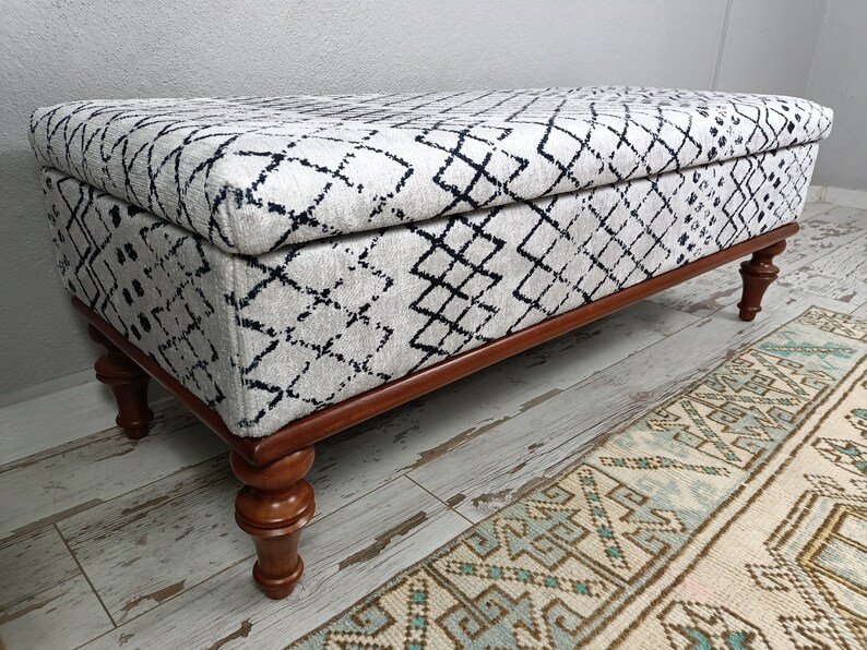 May include: A white and black patterned storage ottoman with a wooden frame and turned legs. The ottoman is sitting on a light wood floor.