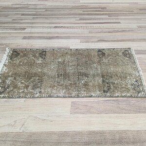 May include: A vintage-style rug with a faded, distressed look. The rug has a light brown background with a subtle pattern of darker brown and green hues. The rug is made of a soft, woven material and has a fringed edge.
