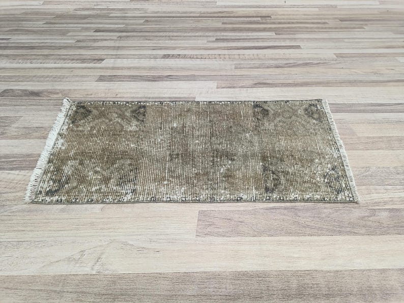 May include: A vintage-style rug with a faded, distressed look. The rug has a light brown background with a subtle pattern of darker brown and green hues. The rug is made of a soft, woven material and has a fringed edge.