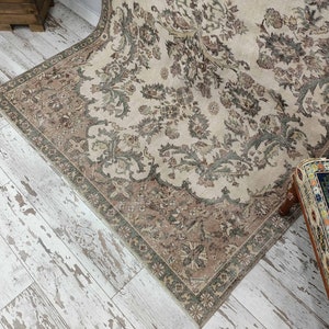 May include: A vintage rug with a beige background and a floral pattern in shades of brown and green. The rug has a fringed border and is laid out on a whitewashed wood floor.
