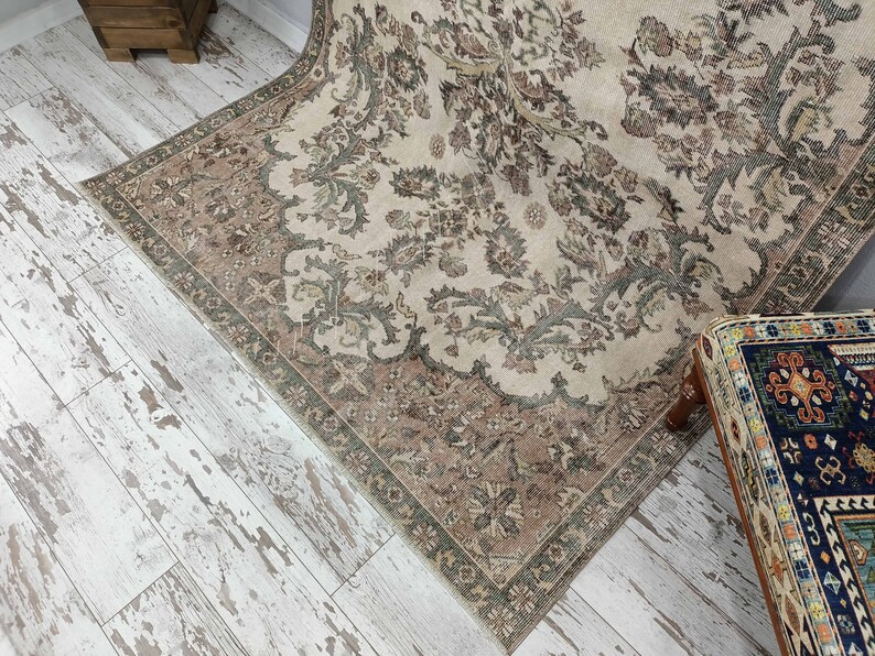 May include: A vintage rug with a beige background and a floral pattern in shades of brown and green. The rug has a fringed border and is laid out on a whitewashed wood floor.