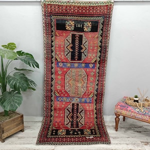May include: A vintage Turkish rug with a red, blue, and black geometric pattern. The rug has a fringe border and is hanging on a white wall.