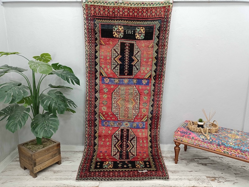 May include: A vintage Turkish rug with a red, blue, and black geometric pattern. The rug has a fringe border and is hanging on a white wall.