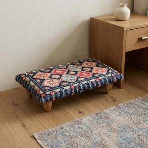 May include: A rectangular footstool featuring a dark blue fabric top with a repeating geometric pattern in shades of red, orange, and light blue. The low-profile stool is supported by four short, rounded wooden legs, providing a stable base.