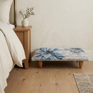 May include: A low rectangular footstool featuring a soft, upholstered top with a large-scale blue and grey floral pattern. The piece is supported by four short, rounded wooden legs, providing a stable base for this functional furniture item.