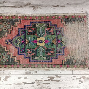 Peut inclure: Un petit tapis vintage avec un motif g&eacute;om&eacute;trique color&eacute; dans des tons de rose, vert, bleu et jaune. Le tapis a un bord effiloch&eacute; et est fait de laine.