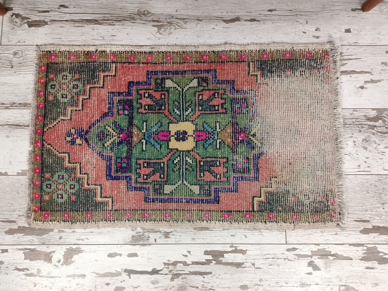 Peut inclure: Un petit tapis vintage avec un motif g&eacute;om&eacute;trique color&eacute; dans des tons de rose, vert, bleu et jaune. Le tapis a un bord effiloch&eacute; et est fait de laine.
