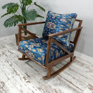 A wooden rocking chair with a blue and white patterned cushion sits in a room with a white wall and a potted plant nearby.