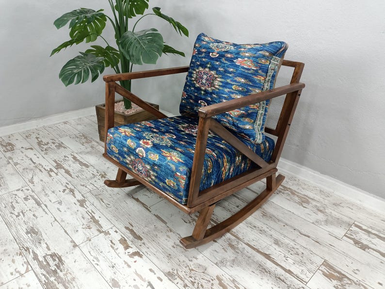 A wooden rocking chair with a blue and white patterned cushion sits in a room with a white wall and a potted plant nearby.
