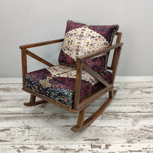 May include: A wooden rocking chair with a patterned cushion and backrest. The cushion and backrest feature a burgundy and cream-colored design. The chair has a dark brown wooden frame and is set on a distressed white wooden floor.