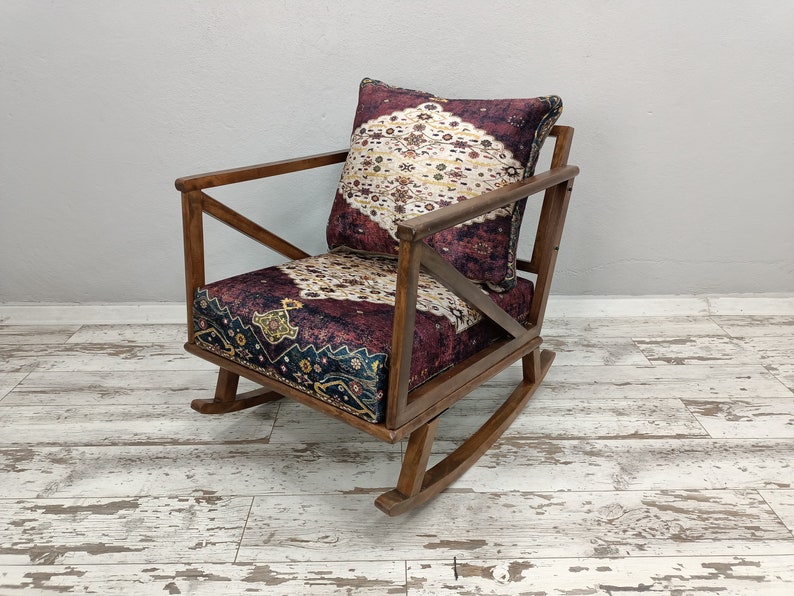 May include: A wooden rocking chair with a patterned cushion and backrest. The cushion and backrest feature a burgundy and cream-colored design. The chair has a dark brown wooden frame and is set on a distressed white wooden floor.