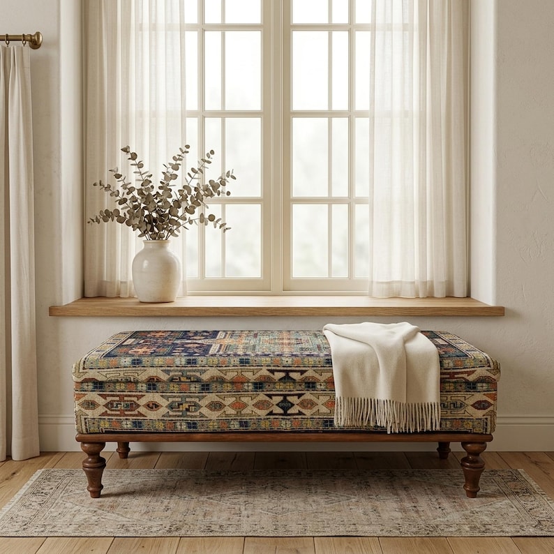 a cozy and inviting living space with a large window, a colorful patterned ottoman, and a vase of greenery on the windowsill.