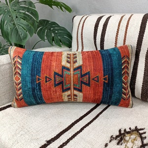 K&ouml;nnte beinhalten: Ein dekoratives Kissen mit einem geometrischen Muster in Orange-, Blau- und Wei&szlig;t&ouml;nen. Das Kissen liegt auf einem wei&szlig;en Sofa mit braunen Streifen.