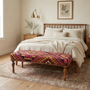 a cozy bedroom with a large bed, a wooden headboard, and a colorful patterned bench. The room has a warm and inviting atmosphere, with a rug on the floor and a framed picture on the wall.