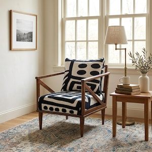 May include: A wooden armchair with a dark brown frame and a white cushion featuring a bold black geometric pattern of circles and squares. The chair has angled armrests and tapered legs, providing a structured seating option for any room.