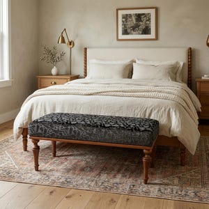 May include: A bedroom with a bed featuring a cream headboard and bedding. A patterned bench is at the foot of the bed. The room includes a wooden bedside table, a framed picture, and a rug with a faded pattern.