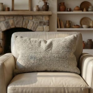 May include: A rectangular, textured, light grey cushion sits on a cream leather armchair. The cushion has a woven appearance and is slightly creased. The background includes a stone fireplace, shelves with pottery, and a mirror.