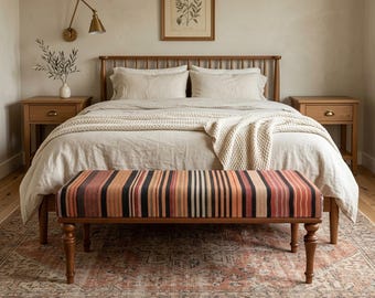 May include: A bedroom scene featuring a wooden bed frame, nightstands, and a bench with a striped pattern. The bed is covered with neutral-colored bedding and a cream-colored throw. A vintage-style rug and artwork complete the cozy aesthetic.