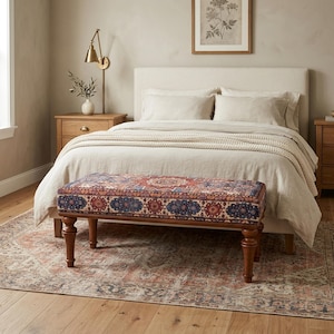 May include: A bedroom scene featuring a bed with neutral-toned bedding and a patterned bench at the foot. The bench has a dark wood frame and a cushion with a red, blue, and cream geometric design. A rug with a similar pattern is on the floor.