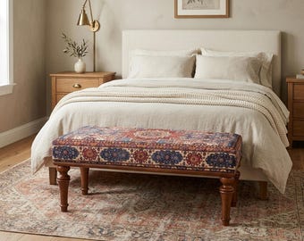 May include: A bedroom scene featuring a bed with neutral-toned bedding and a patterned bench at the foot. The bench has a dark wood frame and a cushion with a red, blue, and cream geometric design. A rug with a similar pattern is on the floor.
