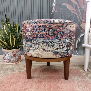 Puede incluir: Un puf redondo con estampado y base y patas de madera. El puf tiene un diseño floral colorido con tonos de rosa, azul y blanco.