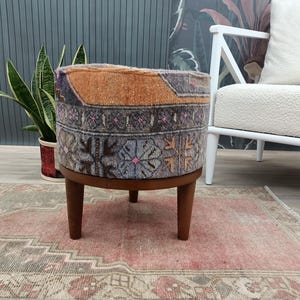Puede incluir: Un otomán redondo con una parte superior de tela estampada en tonos naranja, morado y gris, sobre una base de madera oscura con cuatro patas cortas. El otomán está sobre una alfombra estampada rosa y gris.