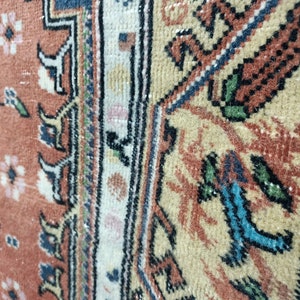 May include: Close-up of a vintage rug with a detailed floral pattern in shades of red, white, and blue. The rug has a fringe border and a worn, faded appearance.