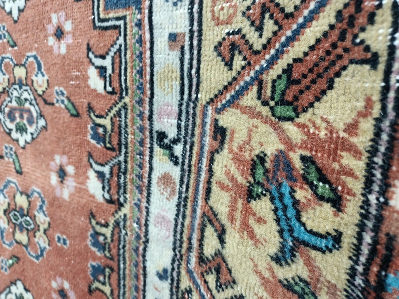 May include: Close-up of a vintage rug with a detailed floral pattern in shades of red, white, and blue. The rug has a fringe border and a worn, faded appearance.