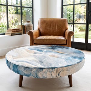 May include: A round coffee table featuring a swirling blue, white, and tan marbled pattern on its surface and sides, supported by short, dark brown wooden legs. The table is positioned in front of a tan leather armchair and a stack of books.