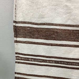 Puede incluir: Primer plano de un textil tejido con un fondo crema y rayas horizontales marrones. Las rayas var&iacute;an en ancho y espaciado, creando un patr&oacute;n texturizado. Los bordes del textil son visibles, mostrando la construcci&oacute;n tejida.