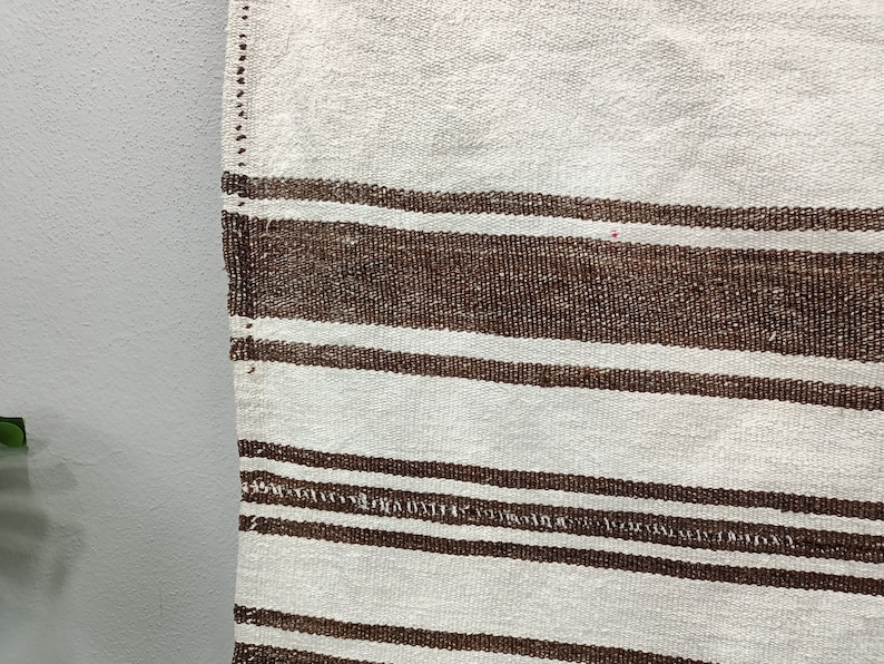 Puede incluir: Primer plano de un textil tejido con un fondo crema y rayas horizontales marrones. Las rayas var&iacute;an en ancho y espaciado, creando un patr&oacute;n texturizado. Los bordes del textil son visibles, mostrando la construcci&oacute;n tejida.