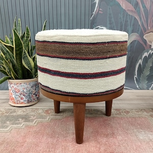 Può includere: Un pouf rotondo a strisce bianche e marroni con gambe in legno. Il pouf è su un tappeto rosa e beige.