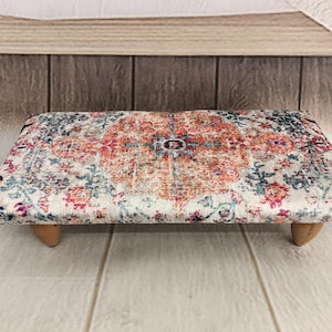 May include: A small, rectangular ottoman with wooden legs. The top is covered in a patterned fabric with shades of pink, orange, blue, and white.