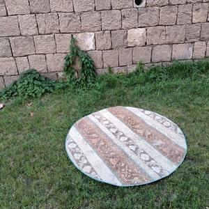 May include: A round, cream-colored rug with brown and blue stripes. The rug is on a green grassy lawn.