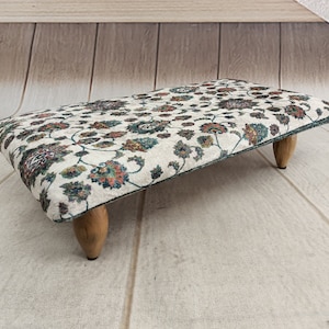 Puede incluir: Un reposapiés blanco con un patrón floral y patas de madera. El reposapiés está hecho de tela y tiene una forma rectangular.