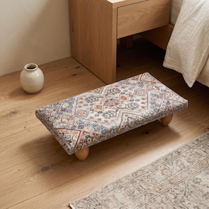 Puede incluir: Un reposapiés rectangular bajo con una parte superior de tela tejida multicolor con un patrón geométrico en tonos azul, naranja y crema. El reposapiés se apoya sobre cuatro patas cortas y redondeadas de madera clara.