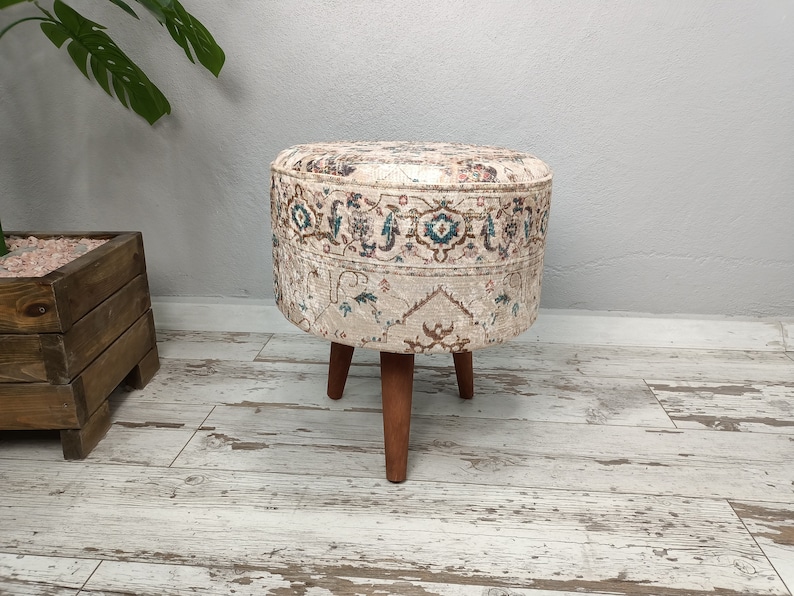 May include: A round, cream-coloured ottoman with a floral pattern and brown wooden legs. The ottoman is sitting on a light wood floor.