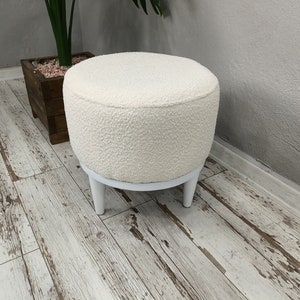Puede incluir: Otomana redonda blanca con una cubierta de tela blanca esponjosa y cuatro patas de madera blancas. La otomana est&aacute; sentada en un suelo de madera blanqueada.