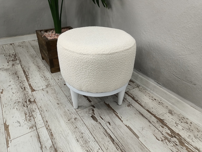 Puede incluir: Otomana redonda blanca con una cubierta de tela blanca esponjosa y cuatro patas de madera blancas. La otomana est&aacute; sentada en un suelo de madera blanqueada.