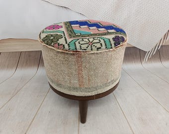 Könnte beinhalten: Ein runder Hocker mit einer Oberseite aus mehrfarbigem Webstoff mit geometrischen und floralen Mustern in Rosa, Blau, Grün und Creme. Die Seiten sind mit strukturiertem beigem Stoff bezogen und stehen auf drei dunkelbraunen Holzbeinen.
