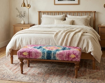 May include: A bedroom with a wooden bed and neutral-toned bedding. A bench with a colourful pink, blue, and yellow pattern sits at the bed's foot. The room is completed with a patterned rug and wooden flooring.