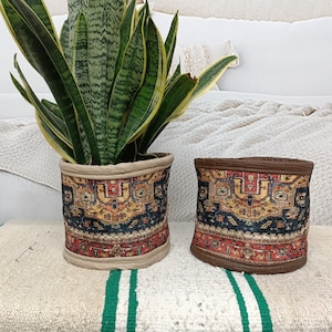 Könnte beinhalten: Zwei zylindrische Pflanztöpfe mit einem gemusterten Design. Die Töpfe sind aus Stoff und haben ein beige, braunes, rotes und blaues Muster. Die Töpfe stehen auf einer weiß-grünen gestreiften Oberfläche.