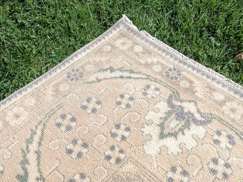 May include: A beige rug with a green and gray floral pattern. The rug has a fringe border.