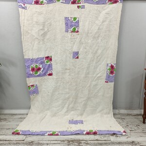 May include: A white quilt with a purple and pink floral pattern. The quilt has a simple design with squares of floral fabric sewn onto a white background.