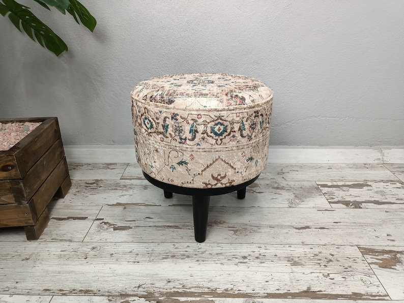 May include: A round ottoman with a cream-coloured fabric cover featuring a traditional Persian-style pattern in shades of brown, blue, and green. The ottoman has four black wooden legs.
