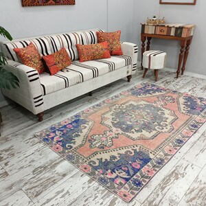 Pode incluir: Um tapete vintage rosa e azul com padr&atilde;o sobre fundo branco. O tapete tem um design floral e est&aacute; colocado sobre um piso de madeira branqueada em frente a um sof&aacute; branco com almofadas listradas.